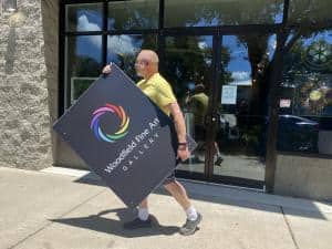 Photo of gallerist and artist Jim Woodfield carrying his logo sign down the street to his new location.